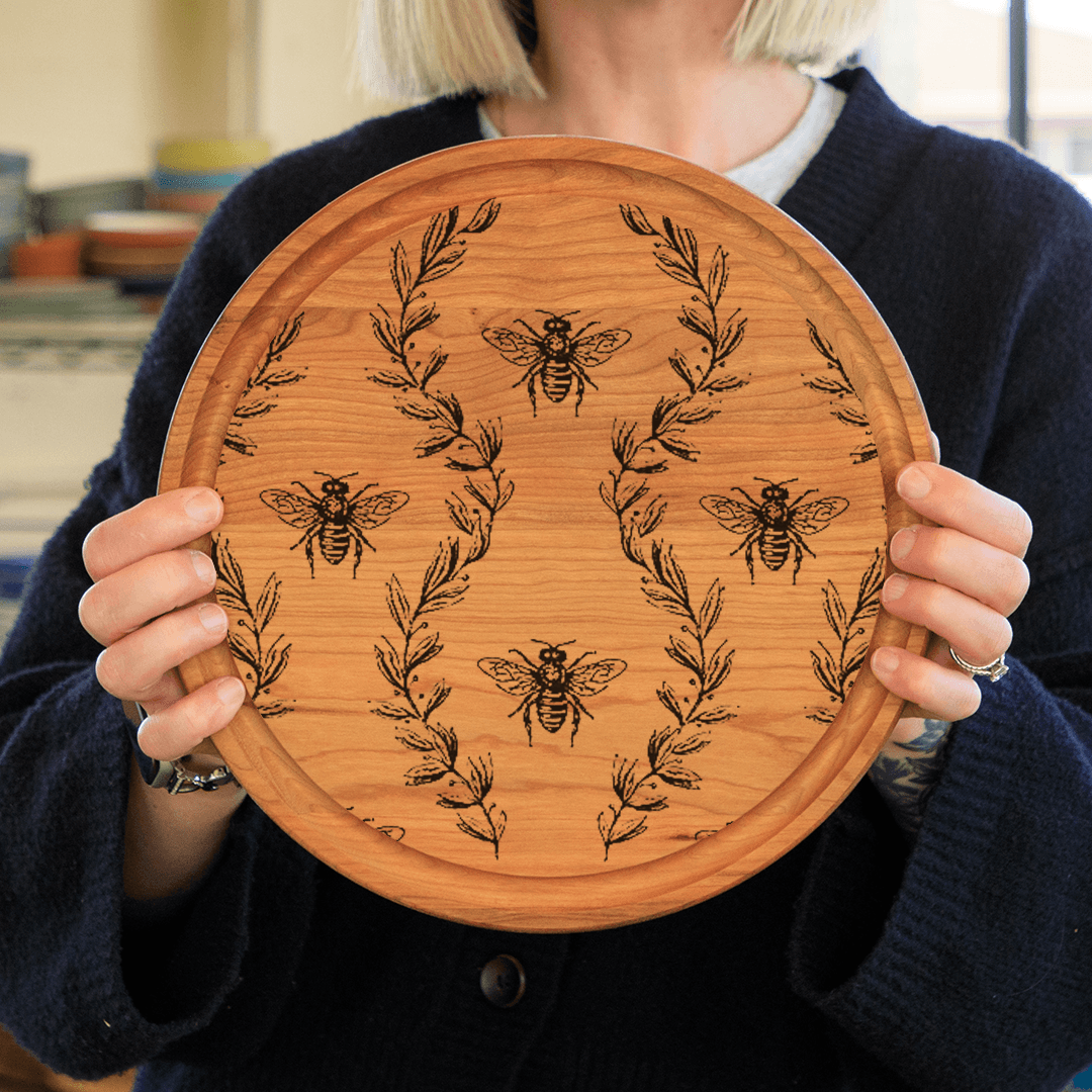 Woman holding a round cherry cutting board with three bees and a laurel wreath wood burned into it